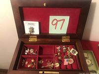 Open wooden jewelry box with red felt interior showing variety of pins and small jewelry items sorted in compartments