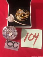 Three ladies pins placed on black velvet with red background. Includes rose crystal pin, bird motif pin, and concentric round pin with glitter. Includes tag labeled 100C Salinas, CA.