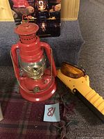 Photo of a red vintage lantern with glass globe, yellow handheld light with amber lens, and wires on carpeted floor