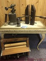 Photo showing antique oil can, wooden carpenter's vice, small metal oil can, and wooden washboard on two-legged table against wooden wall