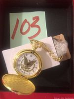 Front view of gold-tone pocket watch with white dial, central eagle graphic, and attached gold-tone chain.