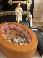 Wooden encased icon of Mary and baby Jesus with crystal flower decorations and pink satin lining, two statues behind, a small framed image, religious plaque, and Student Bible visible.