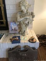 Outdoor woman statue with bird, religious items, and wooden sign on brick surface.
