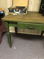 Vintage country style distressed green wooden table with a drawer and metal handle, set against wood-panel wall.