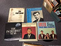 Close-up of a group of vinyl record album covers including S. Kazantzidis, Greek Hit Parade, The Incredible Year, JFK memorial, and Forever on Sunday soundtrack.