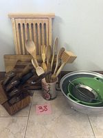 Wooden kitchen utensils in ceramic holder, wooden knife block with knives, wooden cutting boards, and metal colanders.