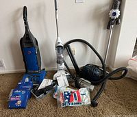 Five vacuums displayed on carpet including upright, canister, and stick styles with accessories and vacuum bags scattered around