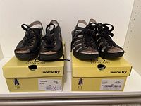 Two pairs of black Fly London wedge sandals on their yellow Fly London shoeboxes, showing front view with laces and insoles