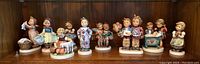 Wide view of all nine Goebel Hummel figurines displayed on a wooden shelf, showing various children in different poses with different accessories.