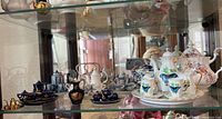 Seven porcelain miniature tea sets and teapots on a glass shelf, multiple styles and colors visible including white, blue and gold sets.