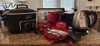 Overview of mixed household small kitchen appliances on kitchen counter including Ninja Cooking System slow cooker, KitchenAid chopper, kettles, and kitchen accessories