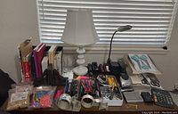 Overview of office desk filled with supplies including lamps, staplers, pens, calculators, scissors, and writing instruments spread out in organizer trays