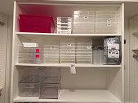 Wide shot of shelving with various sized clear plastic storage containers, including a large opaque pink bin, white perforated baskets, and multiple clear stackable plastic bins with lids.
