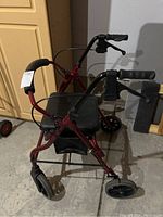 Maroon/red folding mobility walker with black padded seat and hand brakes, showing side angle and front two wheels.