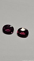 Photo showing two oval polished Umbalite Rhodolite garnet gemstones with deep pink purple color on a white background.