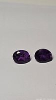 Photo showing two deep purple oval faceted amethyst gemstones placed side by side on a neutral background