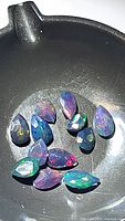 Photo showing eleven pear-shaped calibrated Ethiopian black opal gemstones in a container with vivid rainbow color play under light.