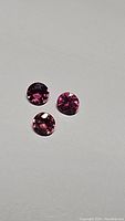 Three bright pink round rhodolite garnet gemstones on a light gray surface under natural lighting.