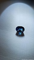 Photo of natural deep blue oval cut sapphire on textured white surface under directed light showing color and facets.