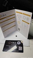 Photo of the GRA certification booklet open showing grading report and certificate number, clear view of loose round moissanite gemstone placed inside.