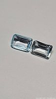 Pair of light sky blue, emerald cut topaz gemstones placed side by side on neutral background, showing their clarity and facets.