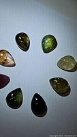 Image showing eight pear-shaped cabochon tourmaline gemstones arranged in a circle on a plain white background. The stones vary in color from green, dark green, amber, pink, and some multicolored with visible inclusions. The cabochon cut gives them a smooth rounded dome surface.