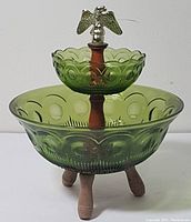 Front view of the green two-tier Coin Glass bowl set with wooden legs and eagle finial on top, showing embossed coin pattern and scalloped edges.