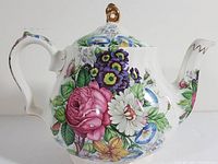 Front angle of white porcelain tea pot with colorful floral design and gold decorative accents on lid and spout tip.