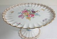 Full view of the white ceramic pedestal cake plate with floral bouquet in center and gold scalloped rim.