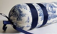 Close-up view of one end of the cylindrical bolster pillow showing cream fabric with blue toile pattern, finished with dark blue ribbon bands and tied blue ribbon end closures.