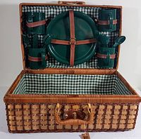 Open wicker picnic basket showing green gingham lined interior with four large green plates, four green mugs, and cutlery each held with leather straps on lid.
