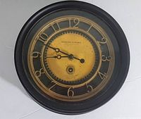 Full frontal view of Sterling & Noble wall clock showing black frame, gold numerals, yellow dial with patina, and clock hands.
