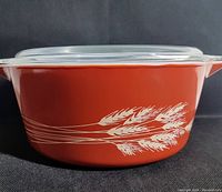Side view of the burnt orange Pyrex Autumn Harvest casserole dish showing the white wheat design and tab handle with the clear glass lid on top.