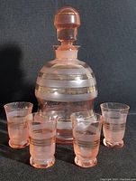 View of the pink glass decanter with gold horizontal stripes and intact stopper shown with five matching pink glasses with gold stripes arranged around it, against a black background.