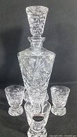 Full view of the footed crystal decanter with stopper and four matching footed shot glasses arranged in front, showing detailed hand cut pinwheel pattern.