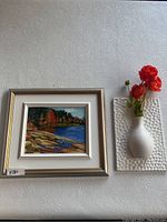Framed landscape painting next to white textured 3D wall decor with artificial flowers on a patterned white wall.