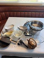 Photo showing all items in lot arranged on table against a window, including ceramic bakeware dishes, glass bowls, large stainless steel pot with strainer and lid, metal meat cleaver, hand press tool, ceramic teapot, and salt and pepper shakers.