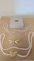 Full top view of two pearl bead necklaces laid out with two pendants (dragonfly and butterfly) and Avon box