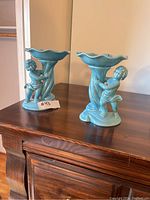 Pair of blue porcelain angel candle holders placed on a wooden surface, showing side view of cherub design and scalloped top bowls.