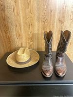 Cowgirl boots and straw hat displayed on dark surface against wooden background.