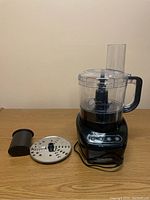 Front view of black food processor with clear work bowl and handle, slicing disc and plastic attachment next to it on table.
