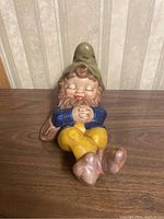 Front view of the ceramic gnome figurine laying down with eyes closed, hands clasped, wearing blue shirt, yellow pants and green hat