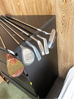 Six golf clubs laid out on black surface showing variety of wooden head and metal irons with wear and brand markings