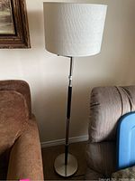Full view of the tall standing lamp placed between two sofas, showing white textured drum shade, black and metal segmented stand, and round metal base.
