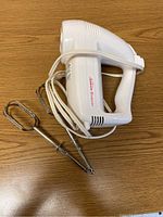 White Sunbeam hand mixer on wood surface with two detachable chrome beaters, power cord wrapped around mixer.