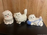 Three white porcelain cat figurines on a black surface against wood background, arranged side by side.