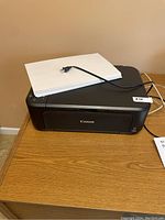 Canon printer placed on a wooden surface with white paper and power cord on top, photographed from front angle