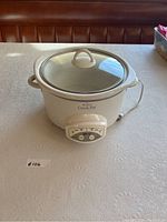 Front angle of white Rival Crock-Pot slow cooker showing the glass lid and side handle placed on textured tablecloth.