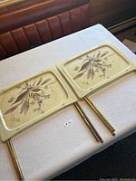 Two vintage rectangular cream-colored dinner trays with brown botanical floral design, set on a white textured tablecloth. Both trays have foldable brass legs attached.