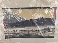 View of the full woodblock print showing Mount Fuji snow capped with a mountain range and trees, with water and small boats in foreground, protected in plastic wrap